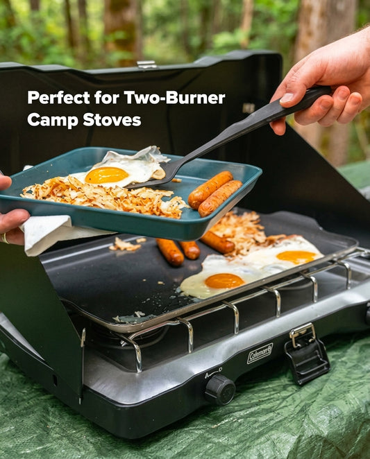 A black Coleman two-burner camp stove outdoors, with a teal frying pan holding eggs and sausage being lifted by a hand using a spatula. The text 'Perfect for Two-Burner Camp Stoves' appears on the left, and a forest background is visible.